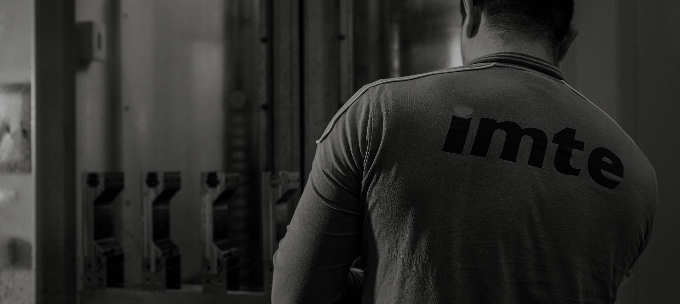 Back view of an IMTE worker wearing branded uniform, standing in front of press brake tooling equipment in a manufacturing environment.